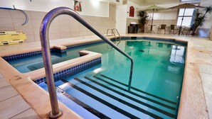 Una piscina interior con una escalera metálica, escalones con azulejos y una superficie de agua azul transparente.