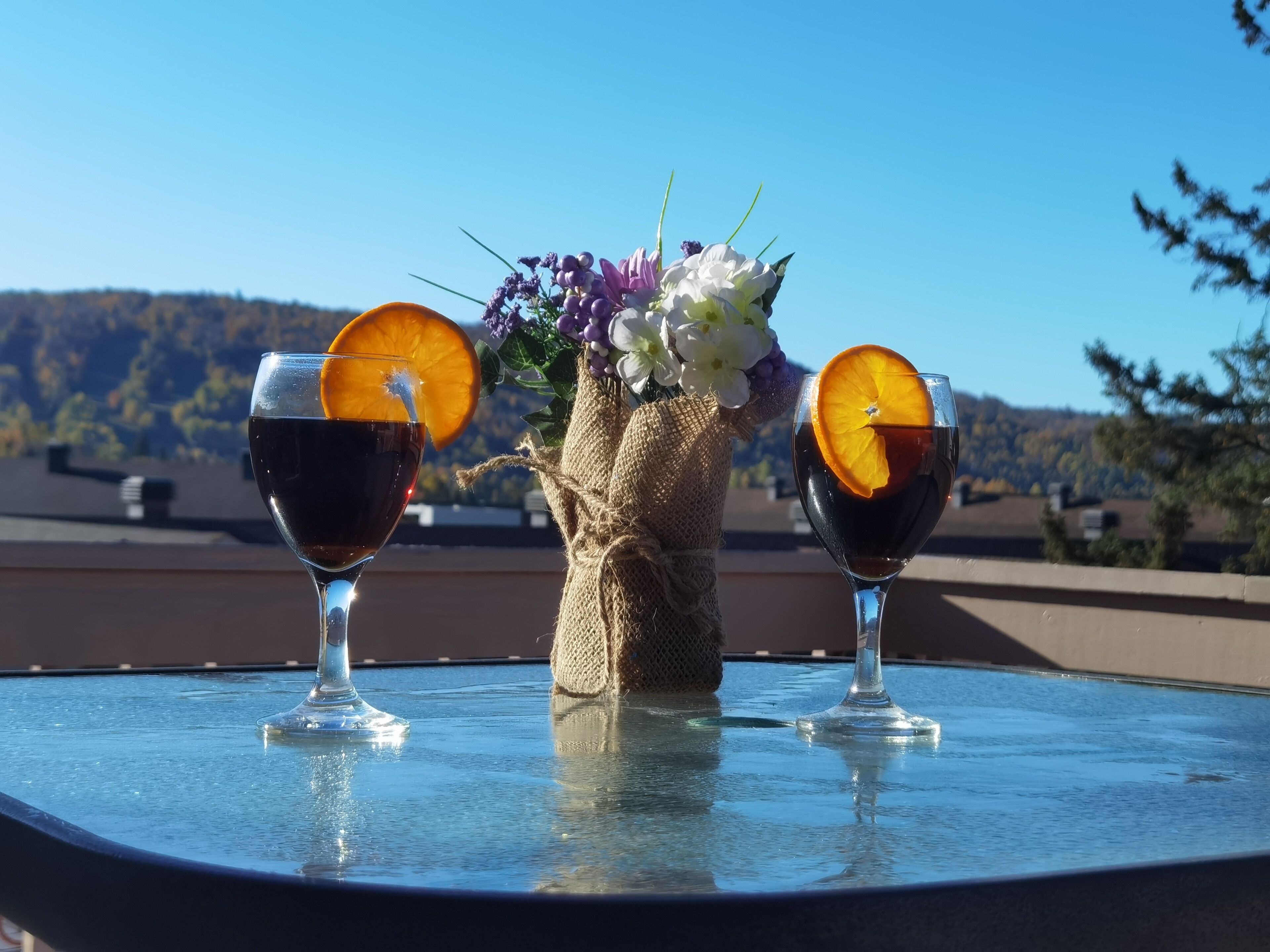To glas med mørk væske og appelsinskiver på et bord med udsigt til et landskab i baggrunden.