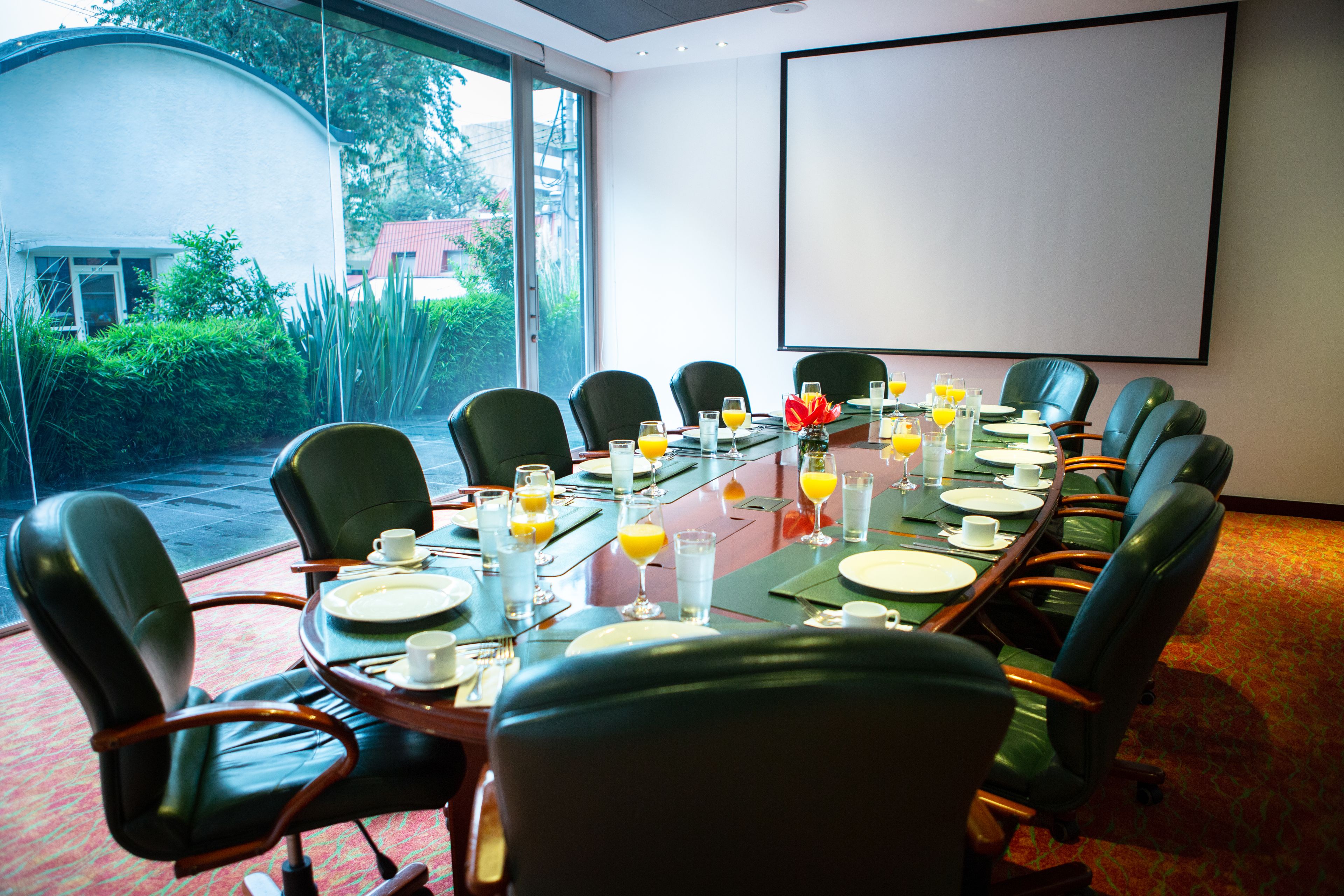 Ein Konferenzraum mit einem langen Tisch, der für ein Meeting gedeckt ist, ausgestattet mit Stühlen, Tellern, Gläsern und einer Projektionsleinwand.
