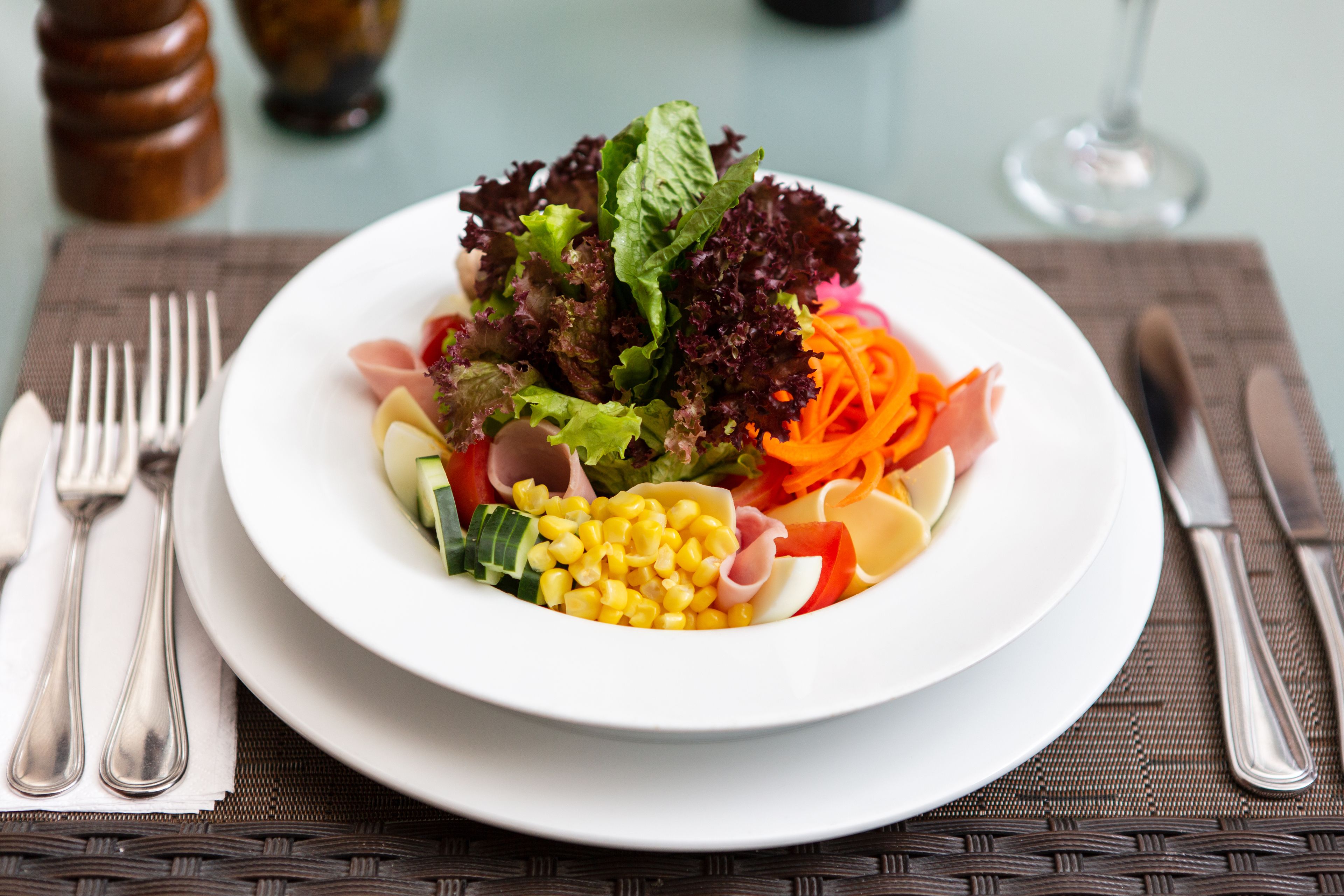 A plate of salad with various vegetables and meats on a white plate, placed on a table with a placemat.