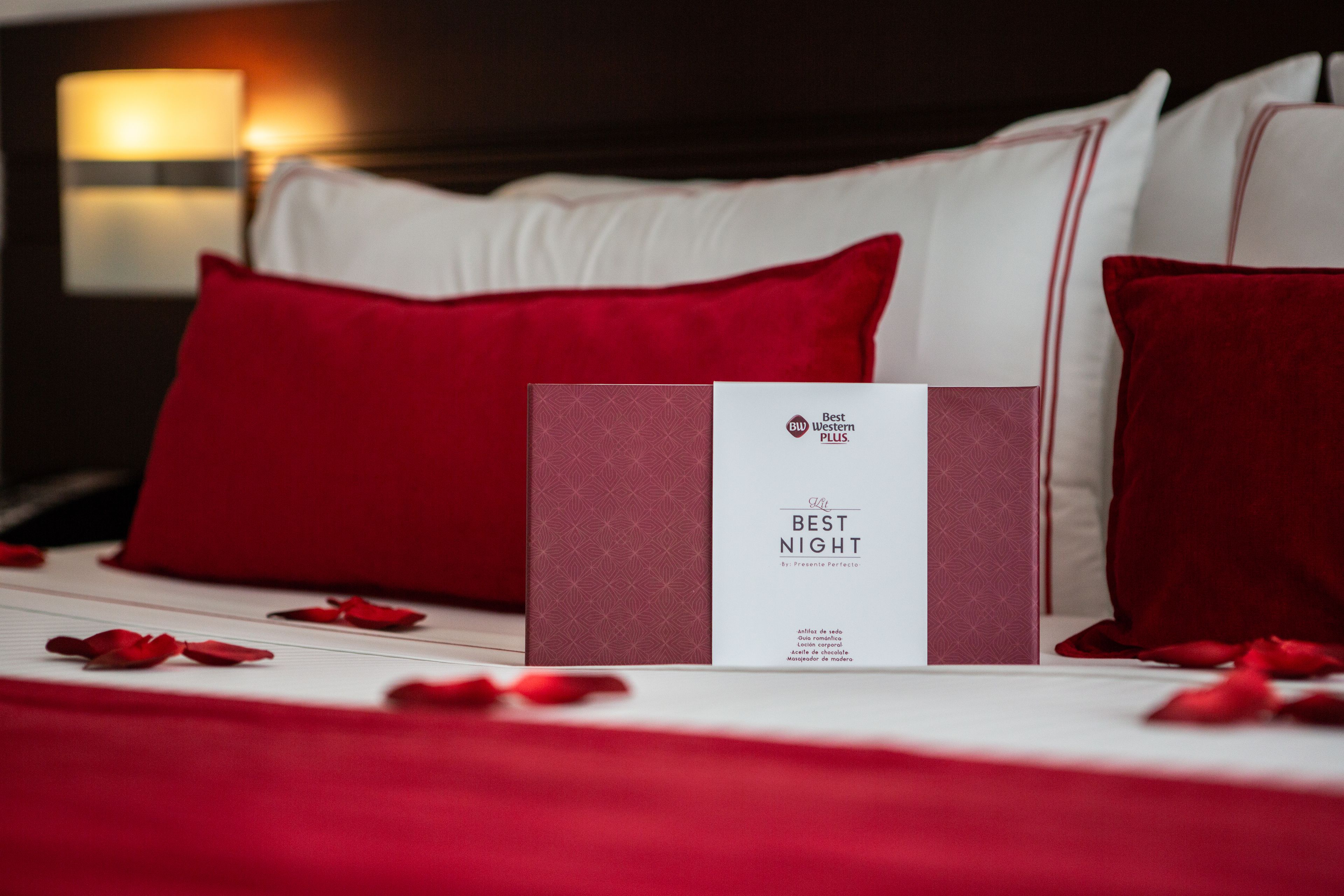 A neatly made bed with red and white pillows, a red rose petal arrangement, and a 'Best Night' brochure on the bed.