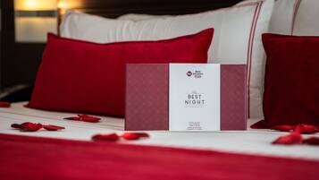 A neatly made bed with red and white pillows, a red rose petal arrangement, and a 'Best Night' brochure on the bed.