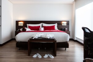 Una habitación de hotel moderna con una cama grande, dos mesitas de noche, suelo de madera y una ventana que deja entrar luz natural.