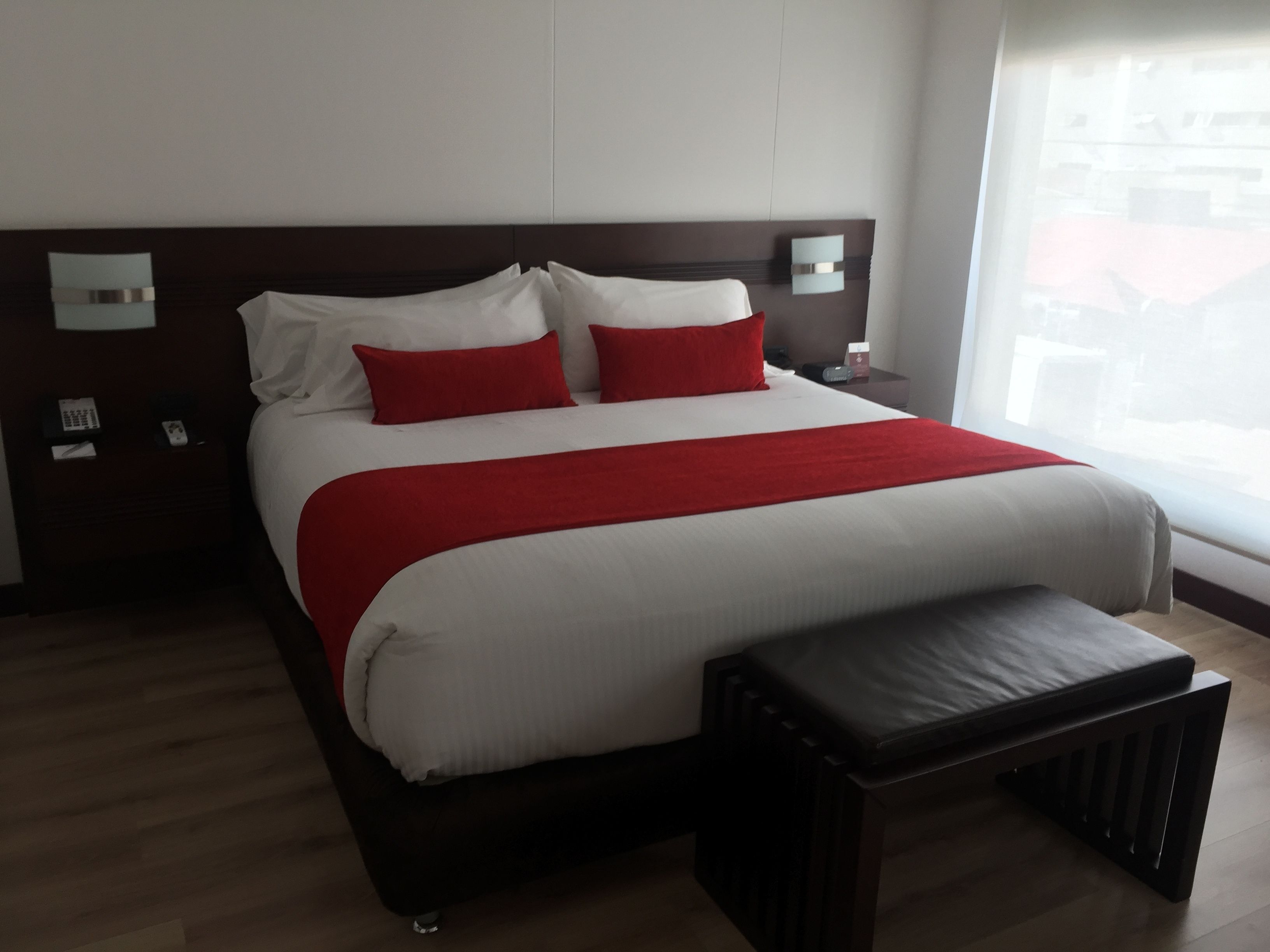 Una habitación de hotel moderna con una cama grande, cabecero de madera, una mesita de noche con lámpara y ventana con vistas a edificios.