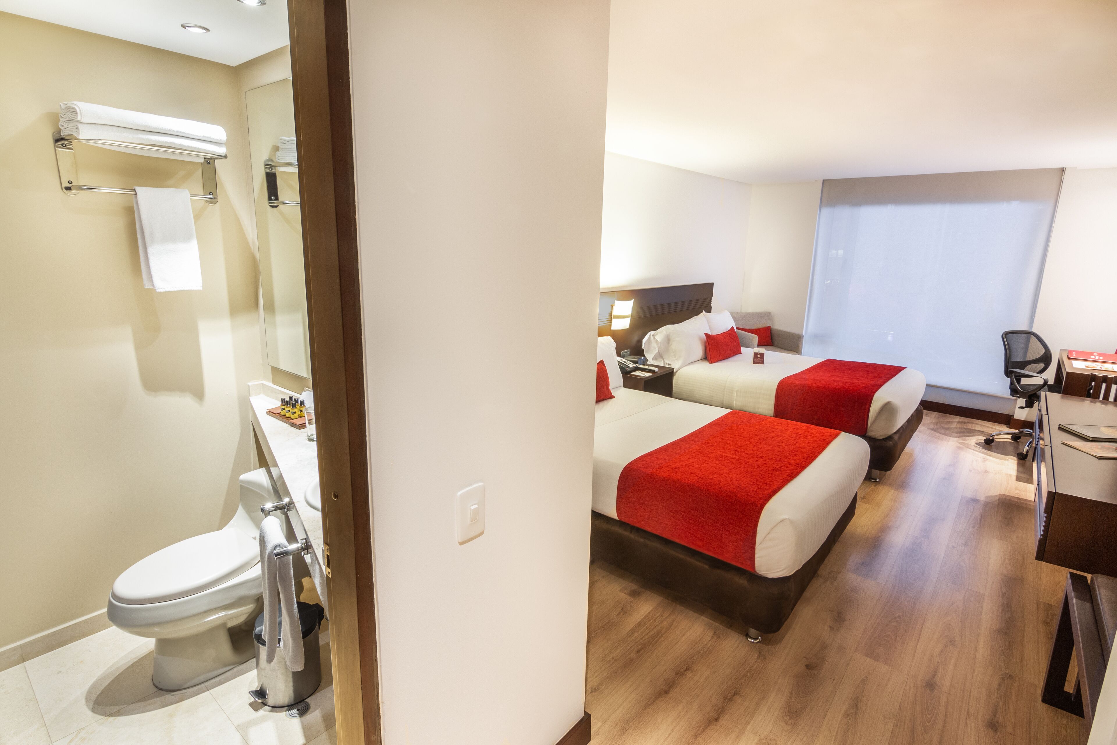 Une chambre d’hôtel avec salle de bain, deux lits avec une literie rouge, un bureau et une fenêtre.