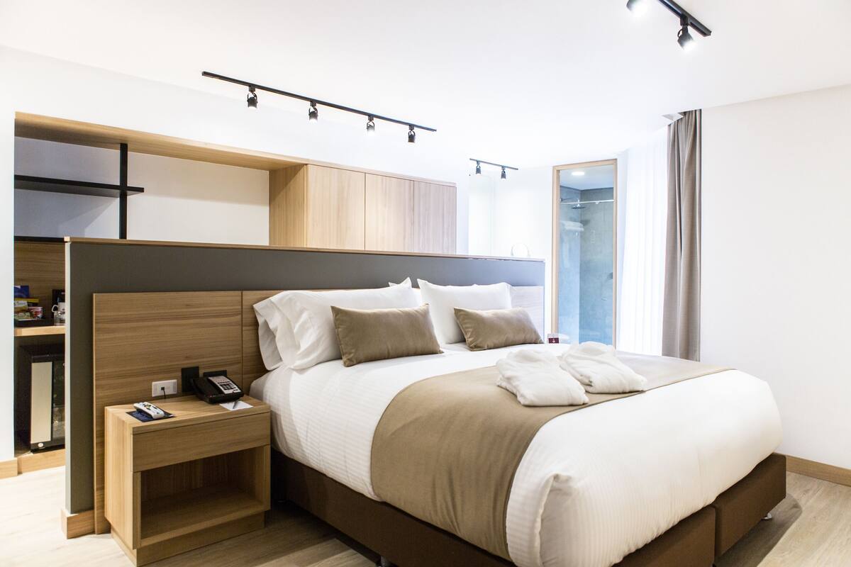 Una habitación de hotel moderna con una cama grande, cabecera de madera, mesita de noche con teléfono y armario empotrado.
