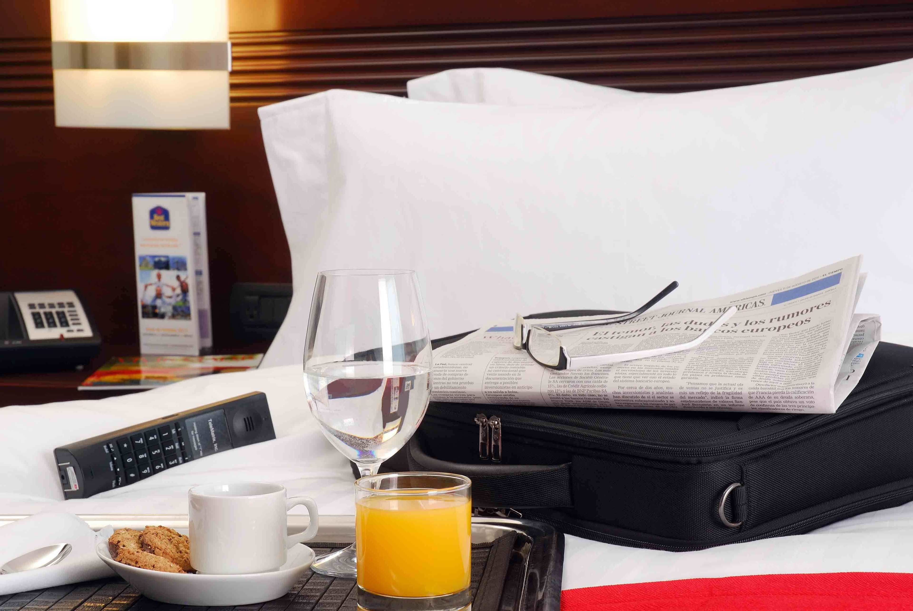A neatly arranged hotel room with a bed, a bedside table with a glass of orange juice, a cup, a plate with cookies, a newspaper, a black suitcase, and a telephone.