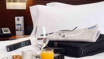 Ett välstädat hotellrum med en säng, ett nattduksbord med ett glas apelsinjuice, en kopp, en tallrik med kakor, en tidning, en svart resväska och en telefon.