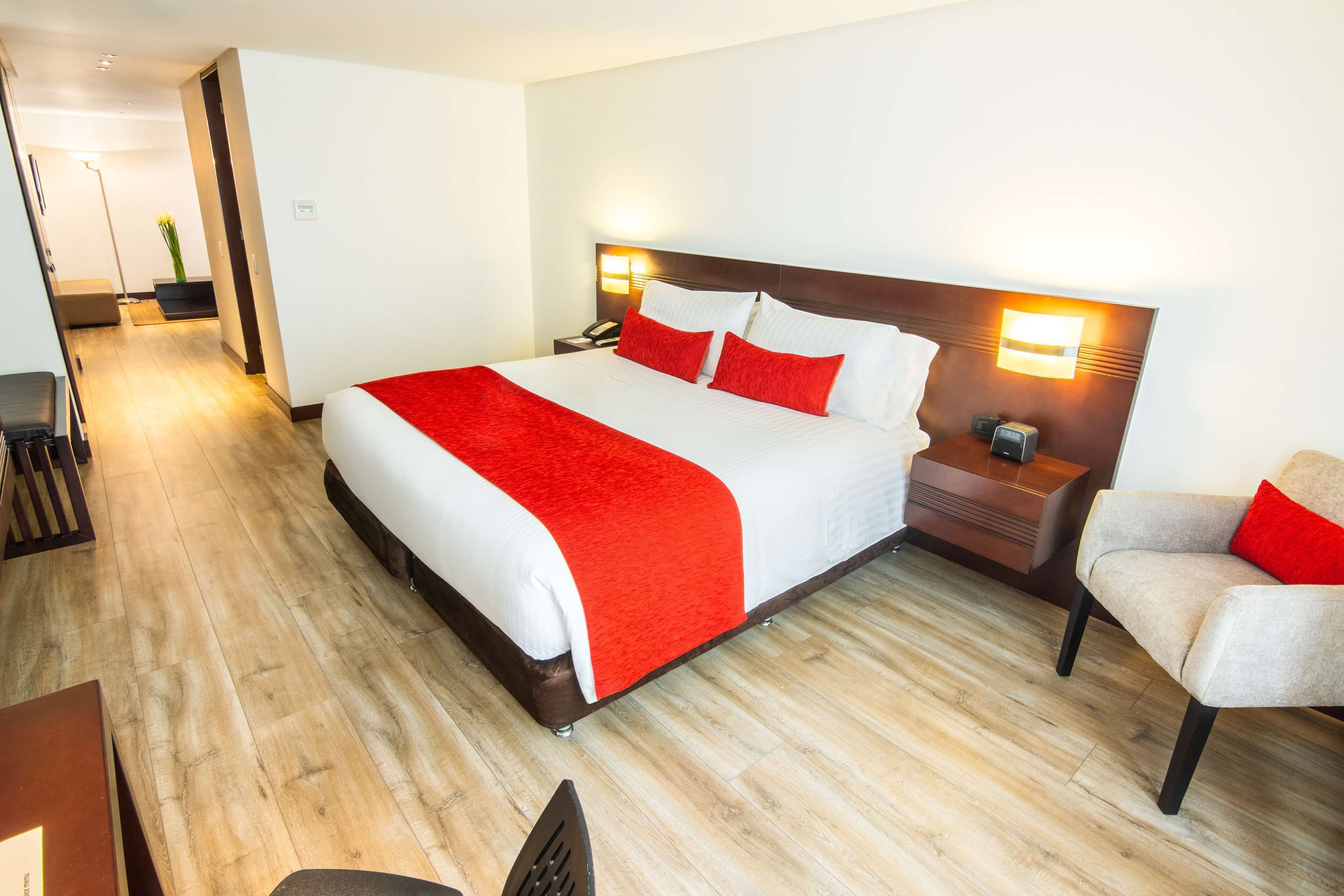 A hotel room with a large bed, a nightstand with a lamp, a chair, and a desk with a chair.