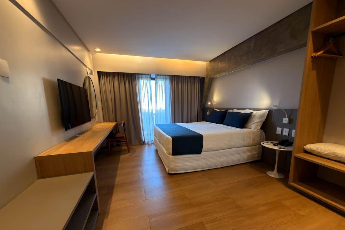 A modern hotel room with a wooden floor, a bed with a blue blanket, a desk with a chair, a TV mounted on the wall, and a window with curtains.