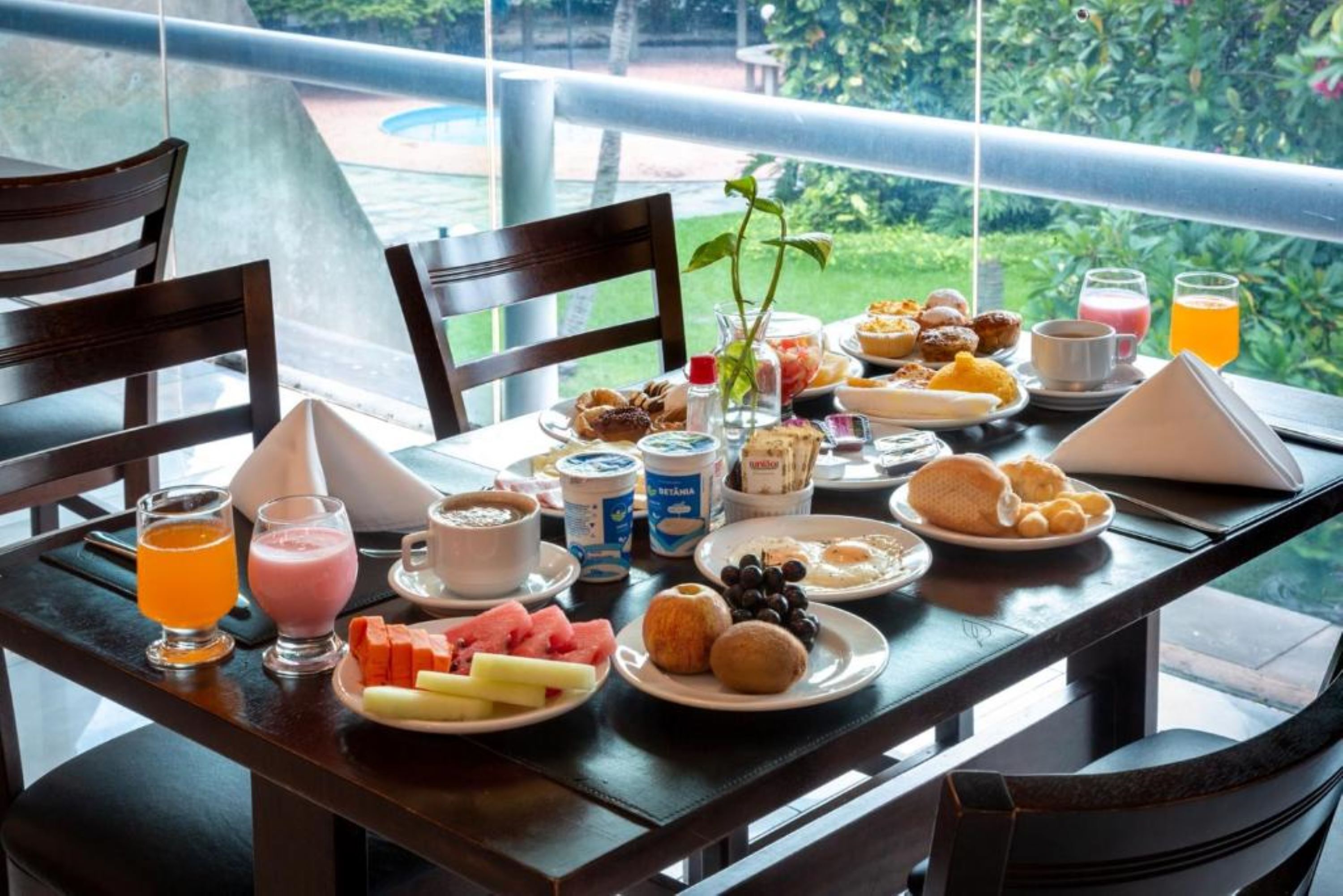 A breakfast table with various foods and drinks, a vase with a plant, and a view of greenery outside.