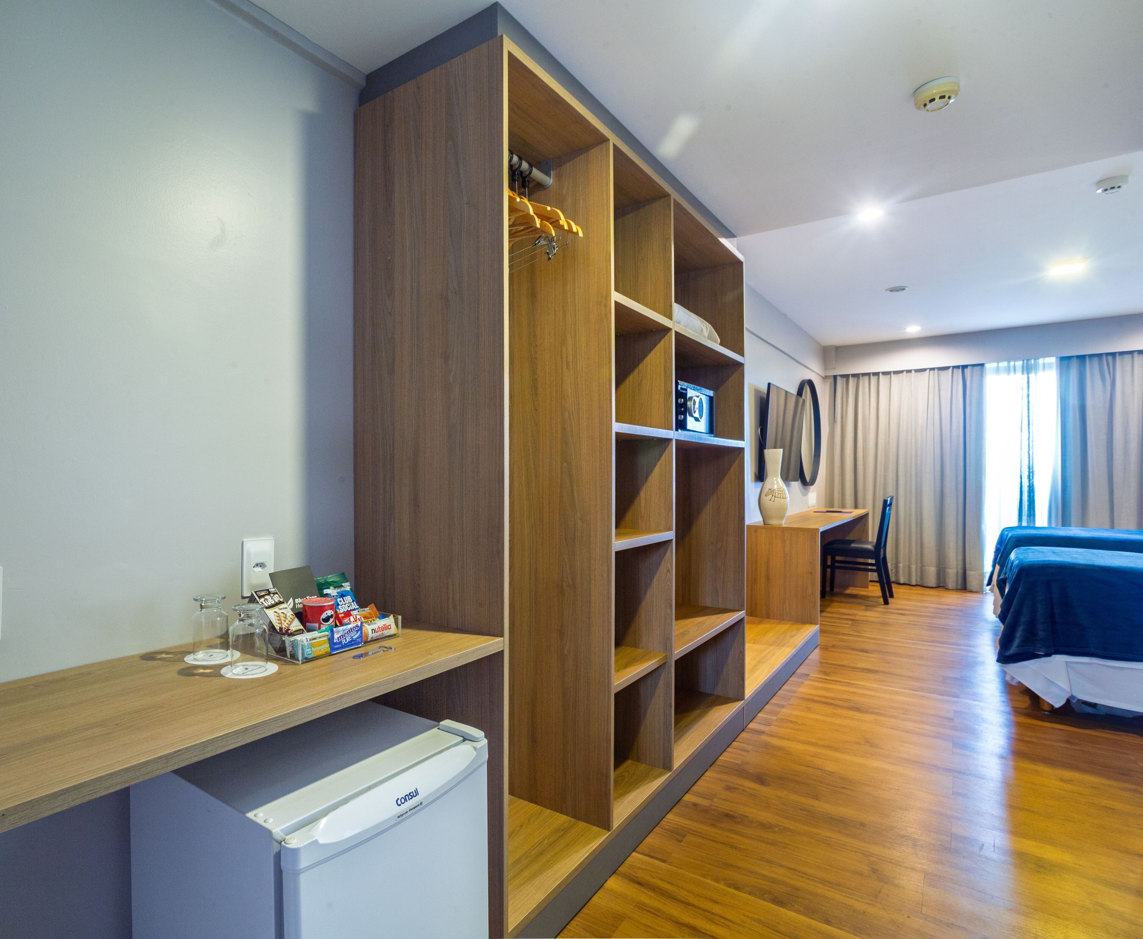 Une chambre d’hôtel avec une armoire en bois, un bureau avec une chaise, un petit réfrigérateur et un lit recouvert d’une couverture bleue.
