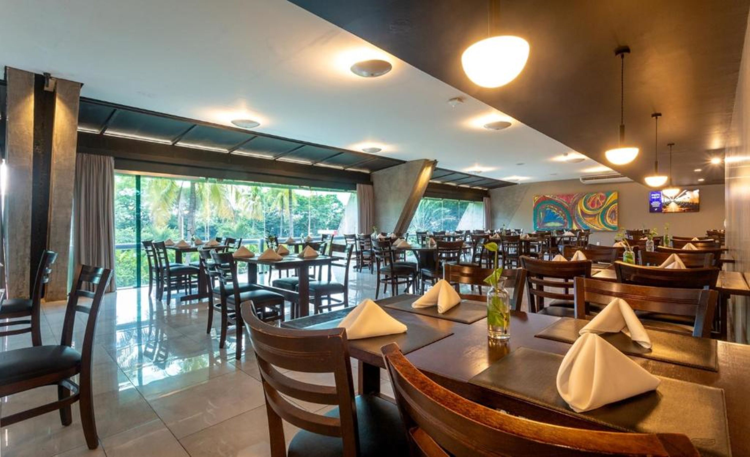 Un restaurante amplio con varias mesas y sillas, grandes ventanales que ofrecen vista a zonas verdes e iluminación de techo moderna.