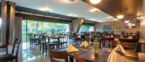 Un restaurante amplio con varias mesas y sillas, grandes ventanales que ofrecen vista a zonas verdes e iluminación de techo moderna.