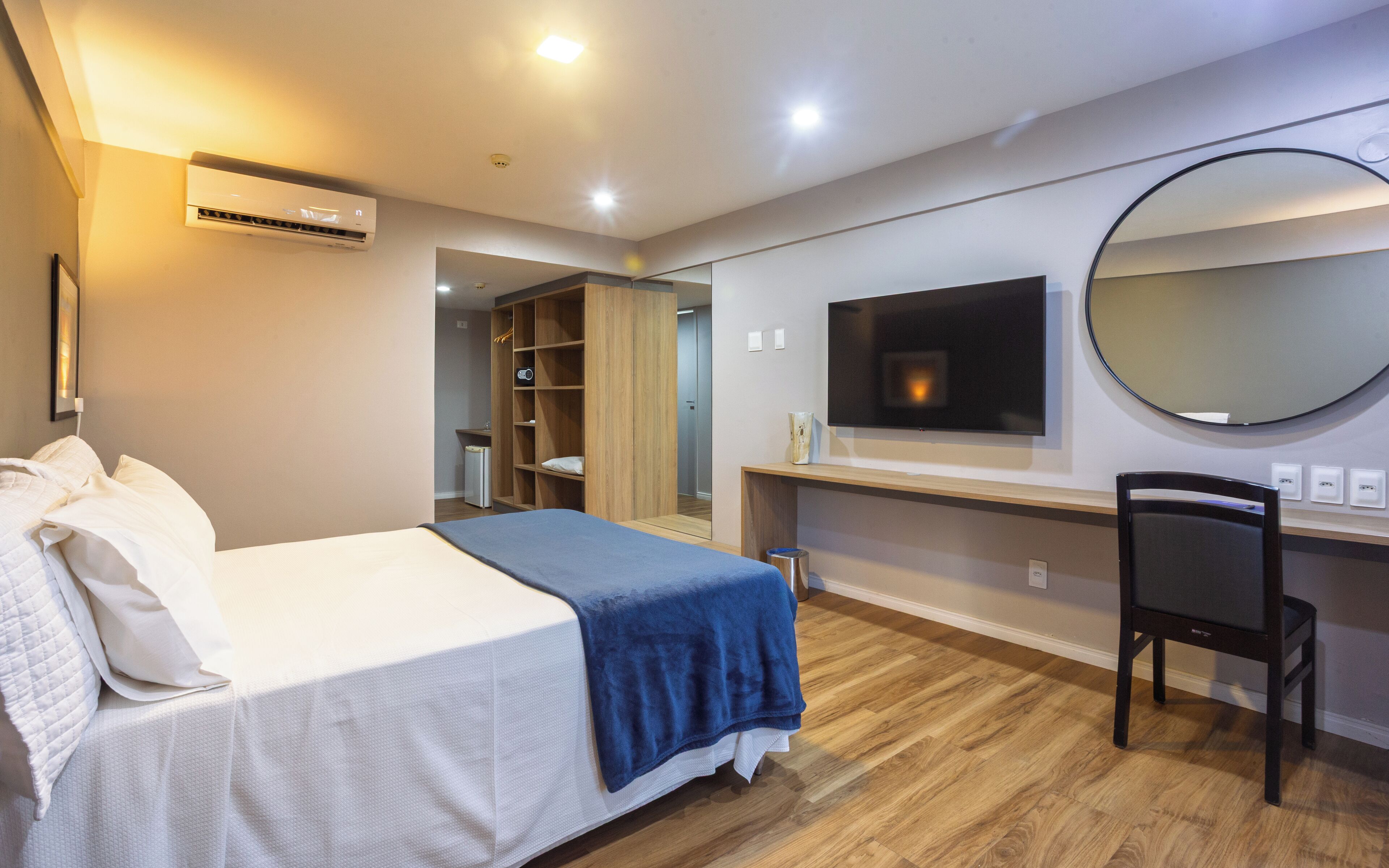 A hotel room with a bed, a desk, a chair, a TV, a mirror, and an air conditioning unit.
