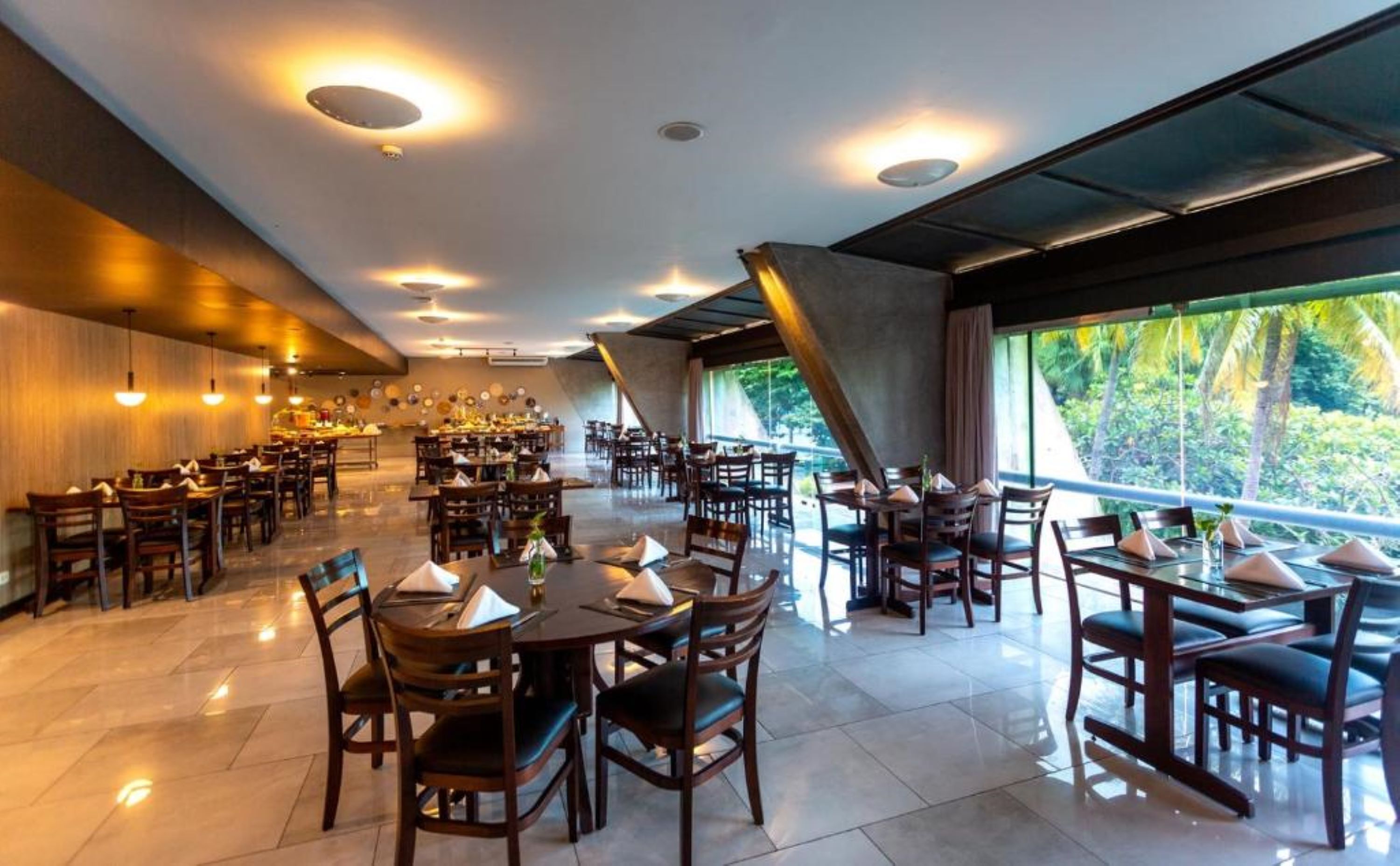 Un restaurant spacieux doté de plusieurs espaces de restauration, de grandes fenêtres offrant une vue sur la verdure et d’un éclairage moderne.