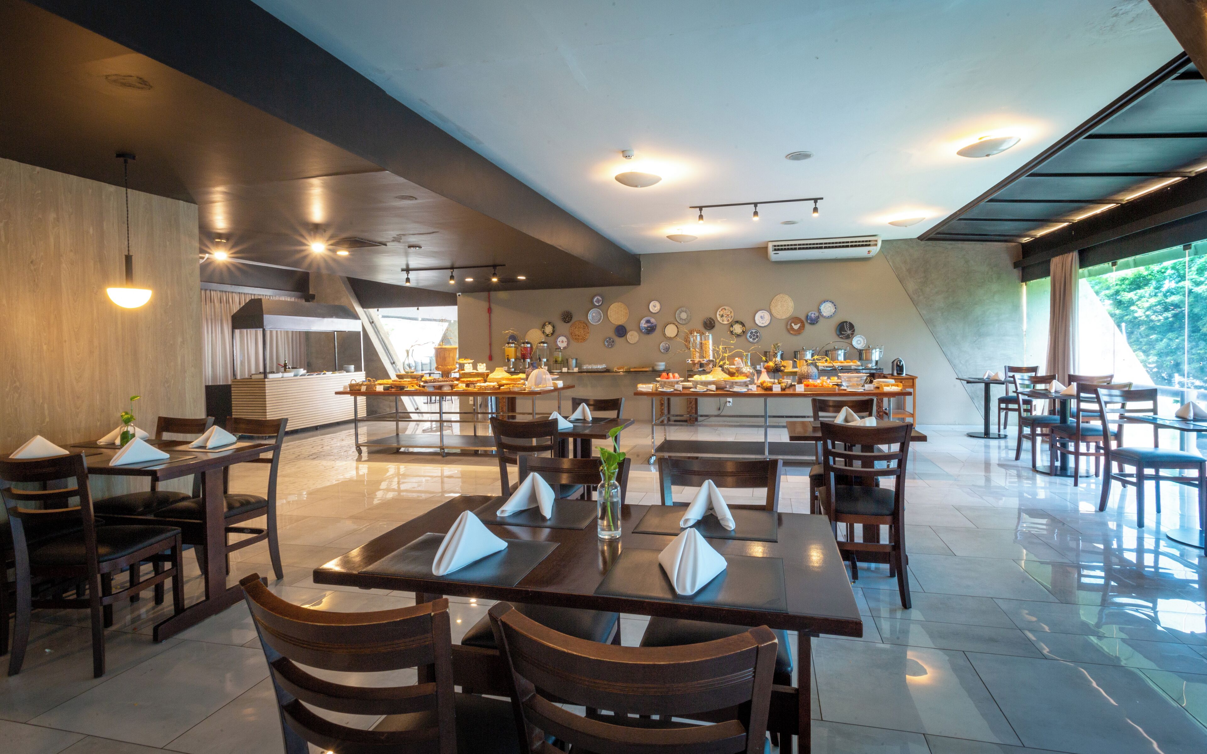A modern dining area with multiple tables and chairs, a kitchen in the background, and a large window providing natural light.