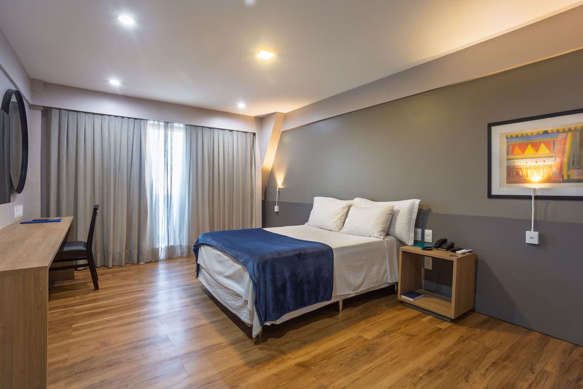 A modern hotel room with a bed, bedside table, desk, chair, and a painting on the wall.