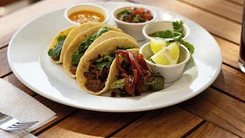 Une assiette contenant trois tacos, un quartier de citron vert et trois petits ramequins de sauces, le tout posé sur une table en bois.