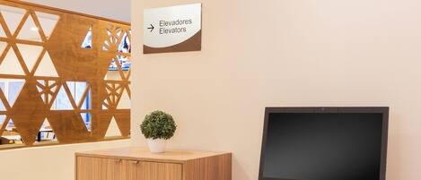A wooden desk with a computer monitor, keyboard, and mouse. A small potted plant is on the desk. A sign above the desk reads "Elevadores Elevators".
