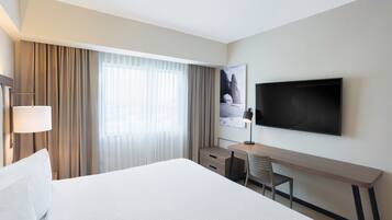 Habitació d'hotel amb un llit gran, una taula de treball, una cadira, un televisor pantalla plana fixat a la paret i una finestra amb cortines.