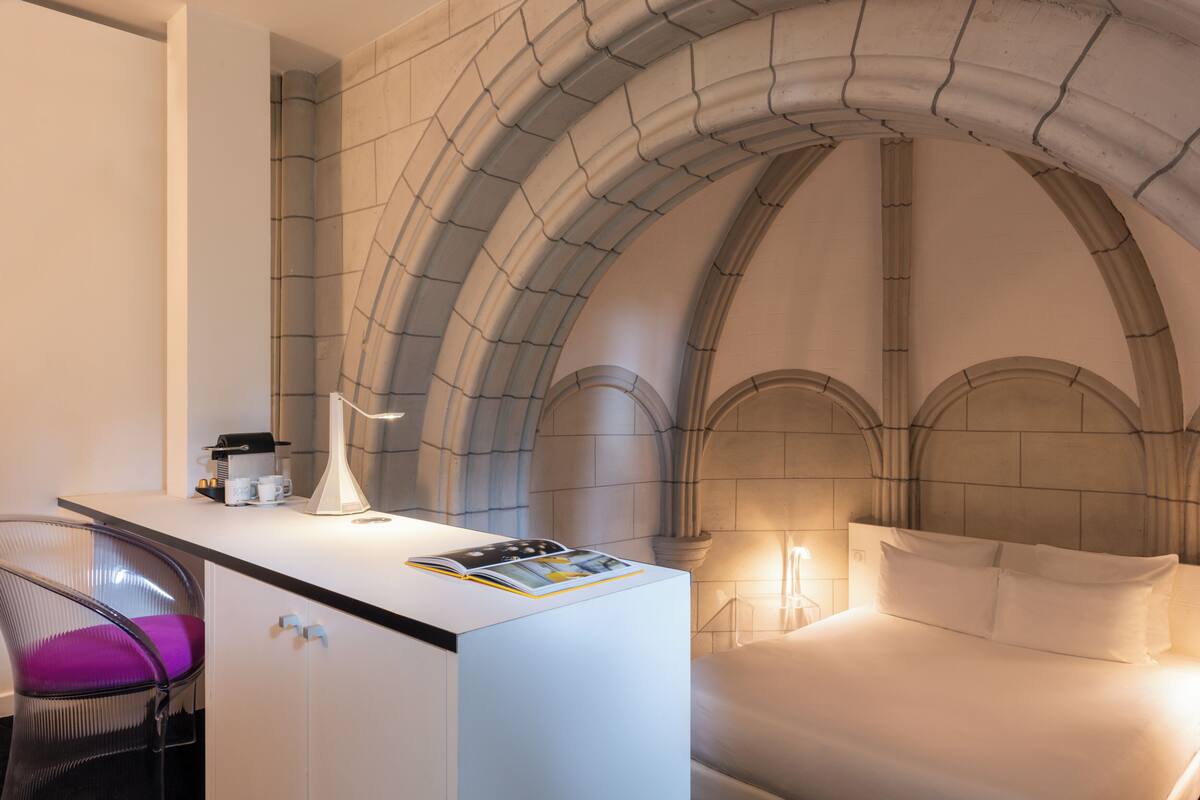 Una habitación de hotel con una cama blanca, un escritorio con lámpara, una silla y una revista.