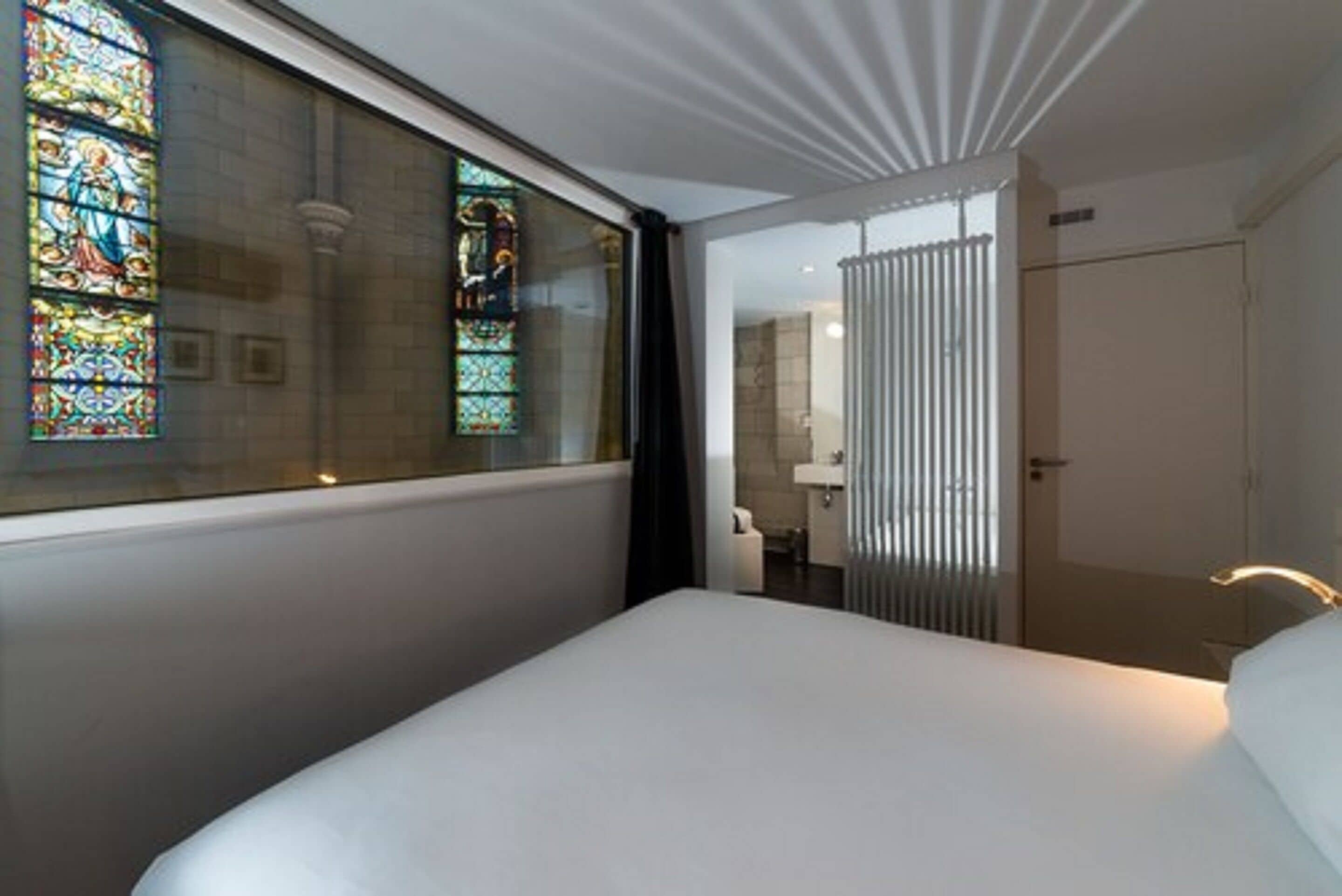 Habitación de hotel con una cama grande, un ventanal con vidrieras, un baño con lavamanos y una puerta.