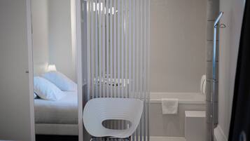 Een moderne badkamer met douche, toilet en wastafel. In de aangrenzende kamer staat een bed met wit beddengoed.