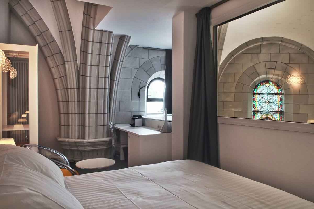 Habitación de hotel moderna con una cama grande, un escritorio pequeño, una silla y un ventanal con vidrieras.
