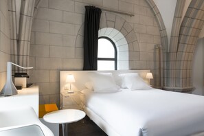 A modern hotel room with a large bed, two bedside tables with lamps, a round table, and a view through an arched window. - Sozo Hotel (Nantes)