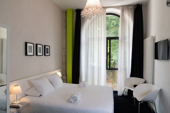 A modern hotel room with a large bed, a bedside table with a lamp, a chair, and a TV.
