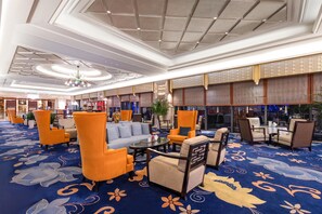 Un hall d’hôtel avec un tapis bleu, des fauteuils orange et beige, un canapé gris et un lustre.