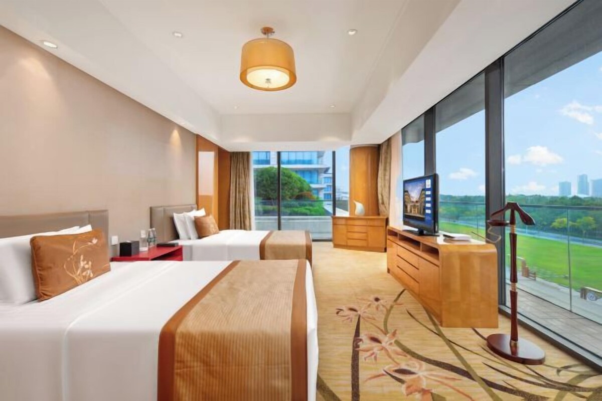 A hotel room with two beds, a TV, a desk, and a large window with a city view.