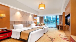 A hotel room with two beds, a desk, a TV, and a large window with a view of buildings and greenery. - WorldHotel Grand Juna Wuxi (Wuxi)
