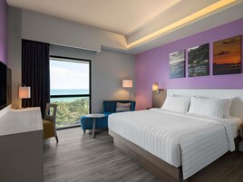 Habitación de hotel con una cama grande, un escritorio, una silla, vista al océano y una pared morada con tres cuadros.