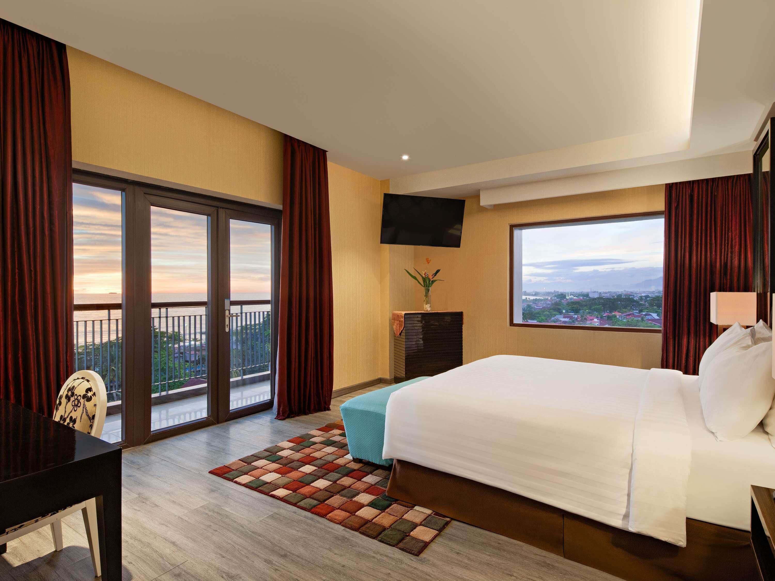 Een hotelkamer met een groot bed, een bureau, een televisie, uitzicht op de stad en een balkon met gordijnen.