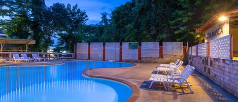 A swimming pool with lounge chairs and a covered area, surrounded by trees and a brick wall.