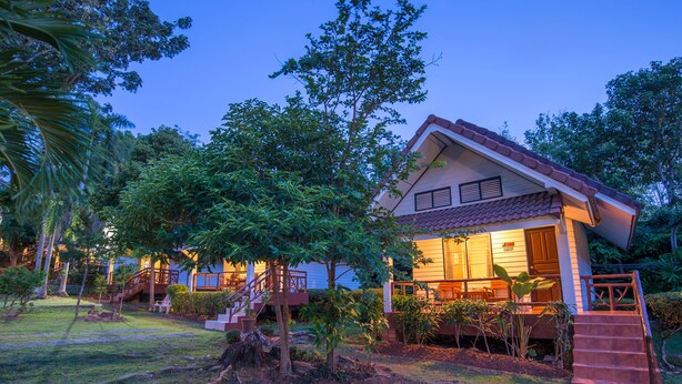 Uma casa de dois andares com telhado vermelho, cercada por vegetação exuberante e um gramado bem cuidado.
