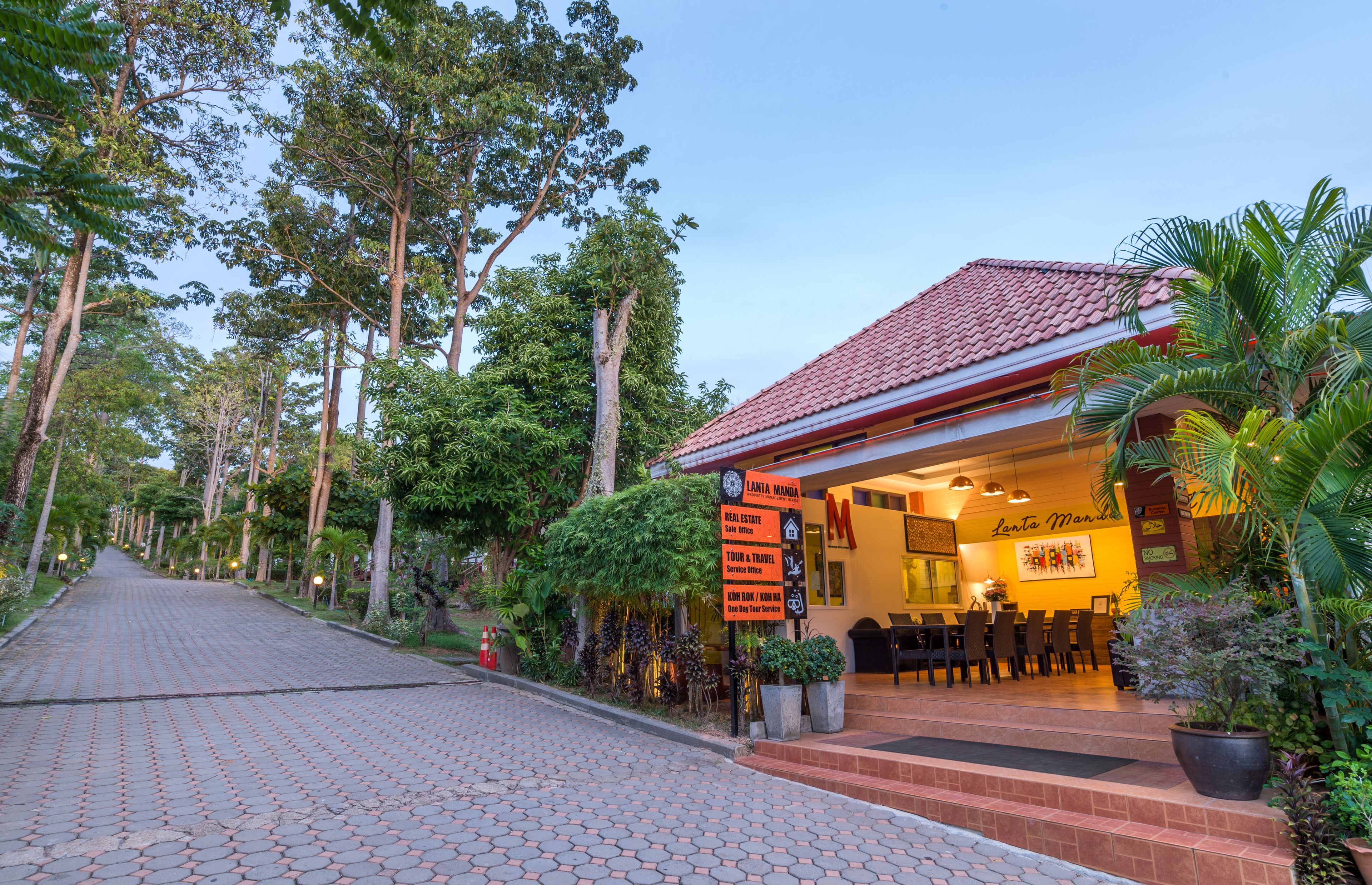 A hotel with a red tiled roof, a reception area with a 'Lanta Manda' sign, and a paved walkway lined with trees.