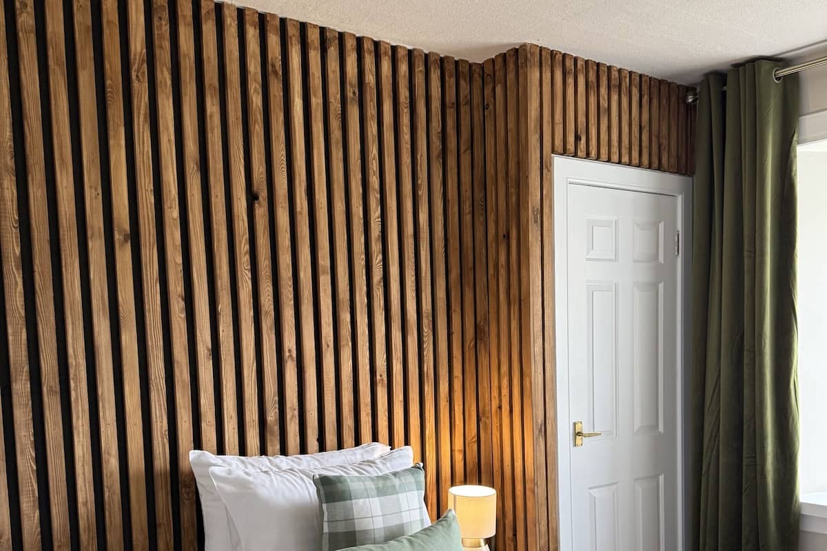 A bedroom with a wooden slat wall, a white door, a bedside lamp, and a bed with white bedding and green pillows.