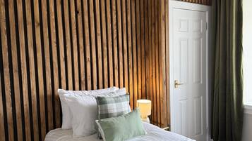 A bedroom with a wooden slat wall, a white door, a bedside lamp, and a bed with white bedding and green pillows.