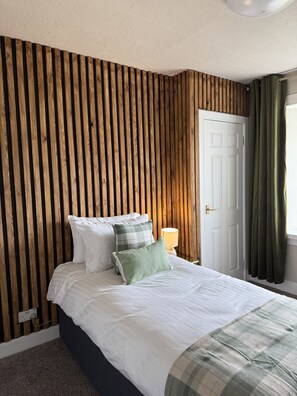 Un dormitorio con pared de listones de madera, una puerta blanca, una lámpara de noche y una cama con ropa de cama blanca y almohadas verdes.