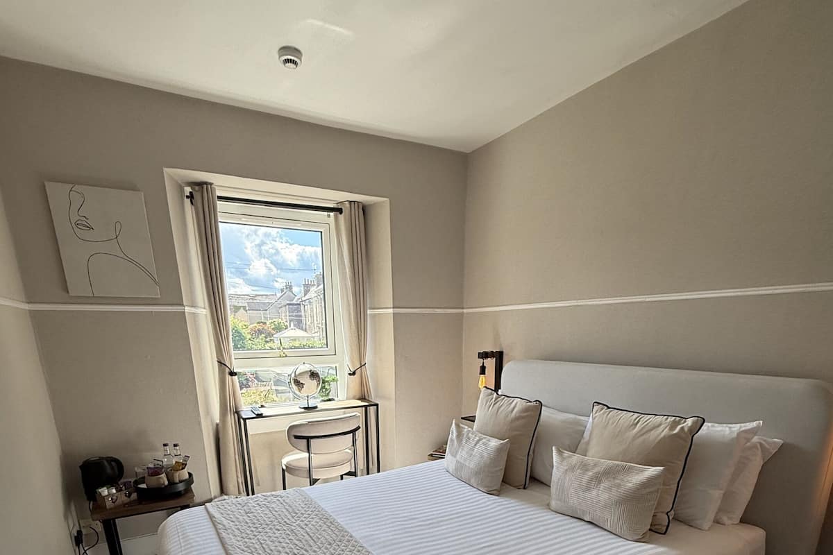 A bedroom with a large bed, a bedside table with a lamp, a window with curtains, and a view of buildings outside.