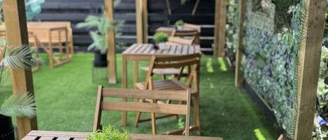 An outdoor patio with wooden tables and chairs, a small potted plant, and a wall covered in greenery.