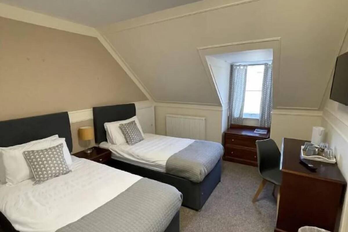 A bedroom with two beds, a desk, a chair, a radiator, and a window with curtains.