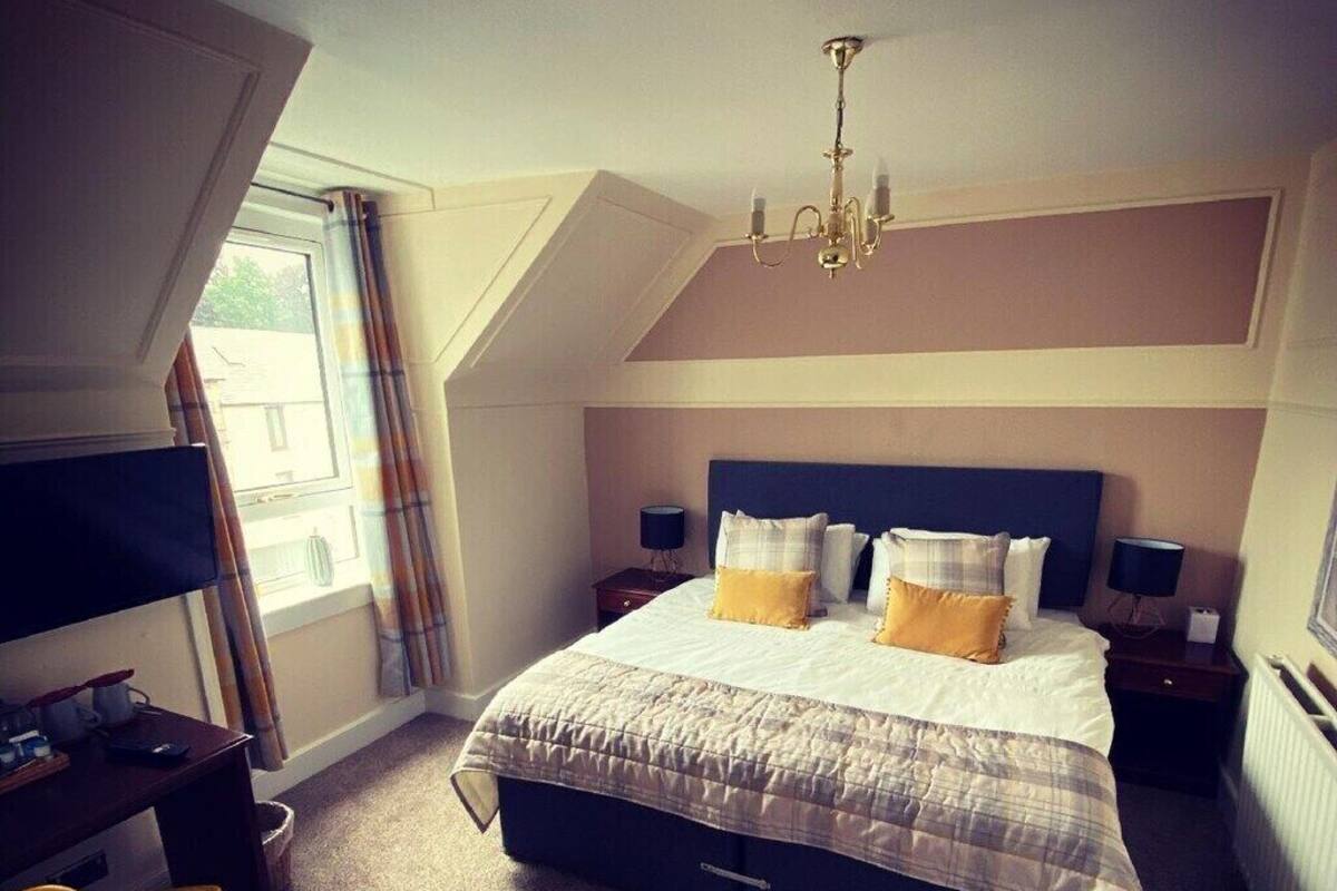 A bedroom with a large bed, a desk, a chair, a chandelier, and a window with curtains.