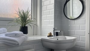 Et badeværelse med en hvid murstensvæg, et rundt spejl, en vask, en potteplante og håndklæder på en radiator.
