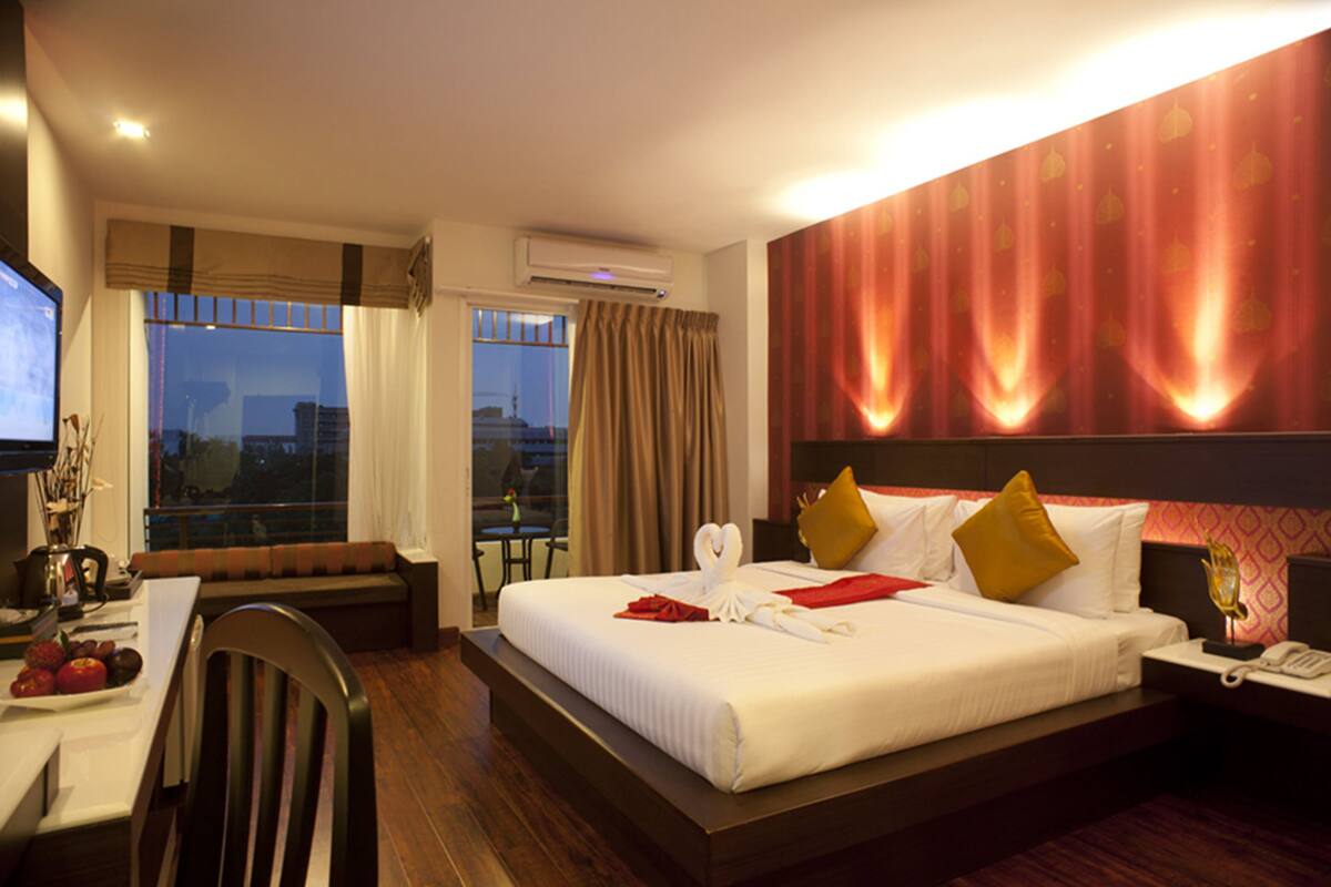 A hotel room with a large bed, a desk with a chair, a dining table with fruit, and a view of the city through the window.