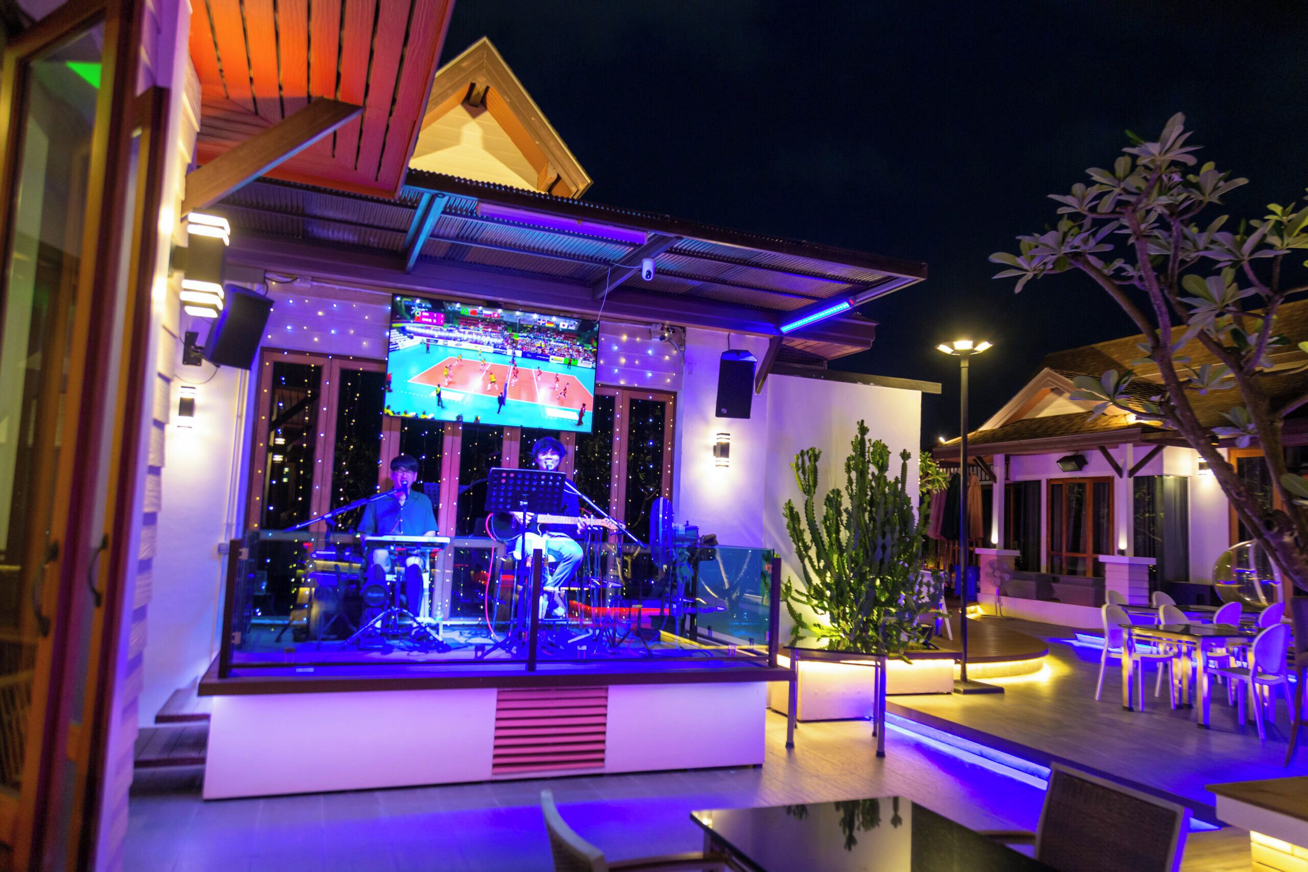 An outdoor bar area with a stage, seating, and a large screen displaying a sports event.
