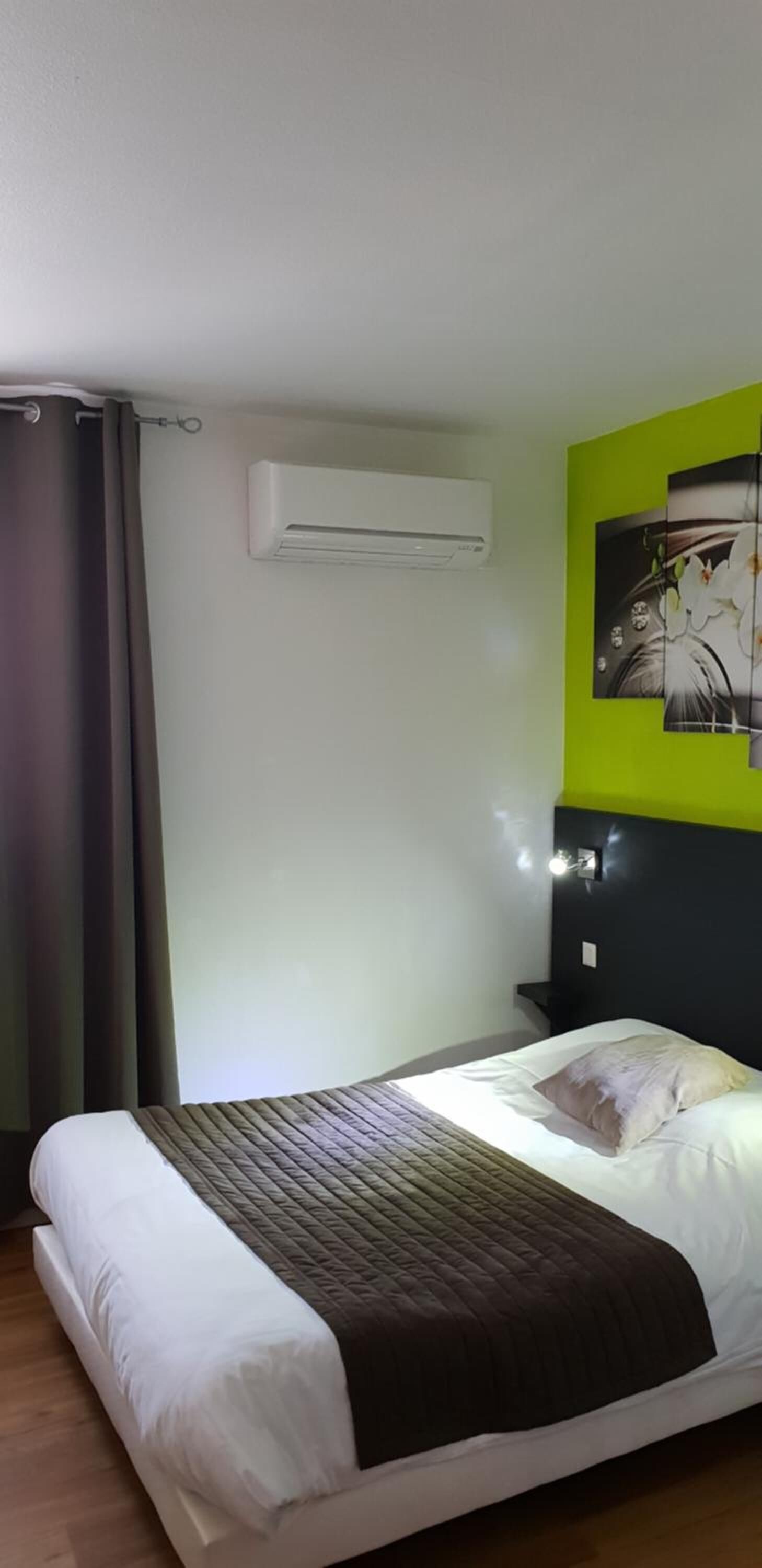 Quarto de hotel com cama, ar condicionado, luminária na parede e uma parede verde decorativa.